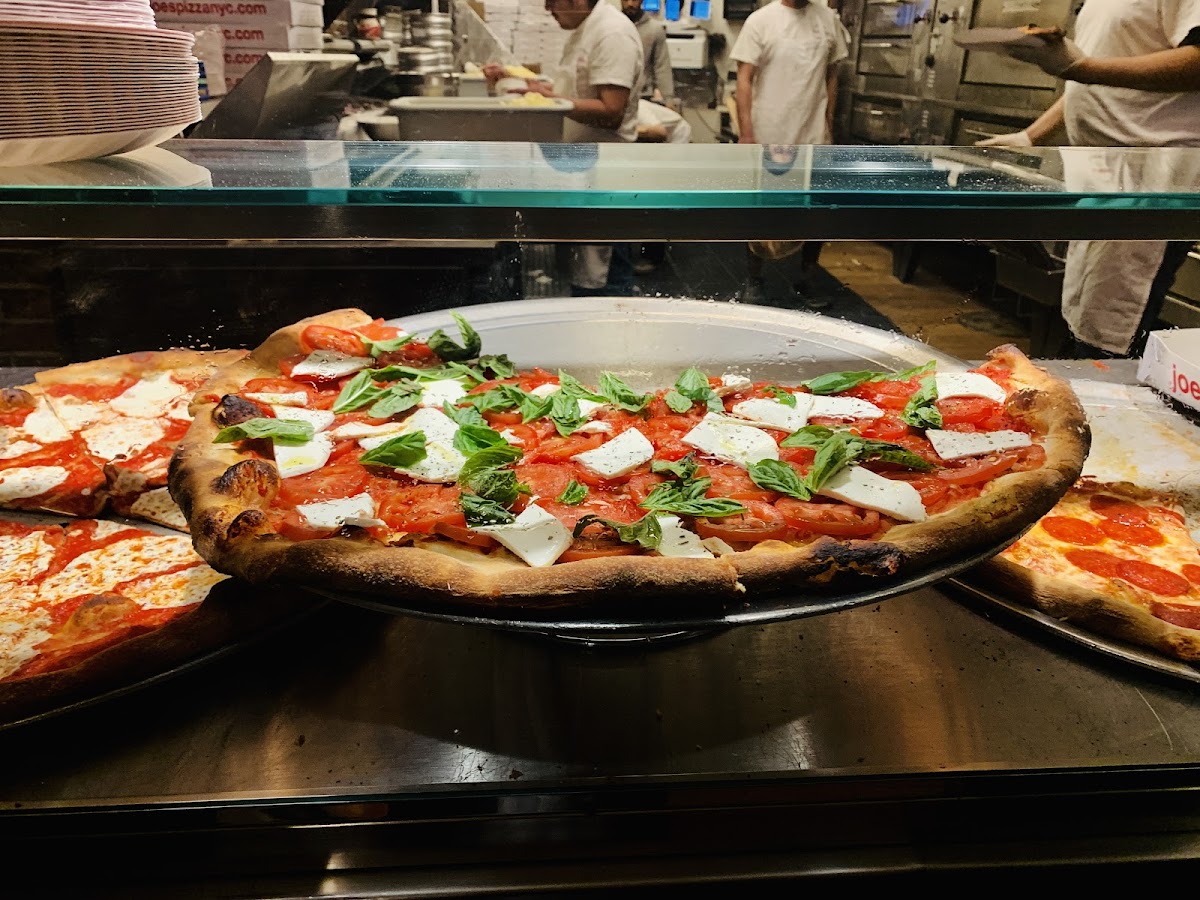 Wide foldable cheese slice on a paper plate at Joe's Pizza on Carmine Street in Greenwich Village, New York, with the storefront counter visible behind.